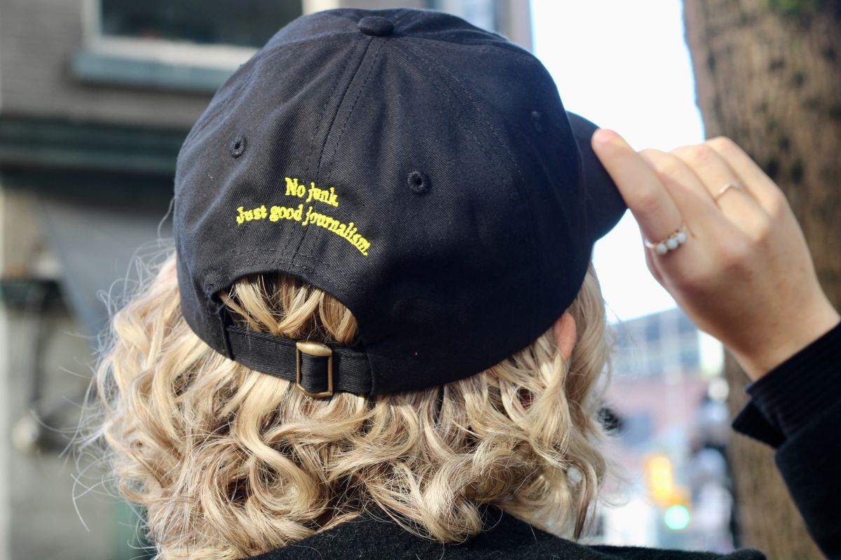 A person with short curly blond hair has her back turned to the camera. She is tipping the bill of the black ball cap she is wearing. In yellow text across the back, it reads &ldquo;No junk. Just good journalism.&rdquo;