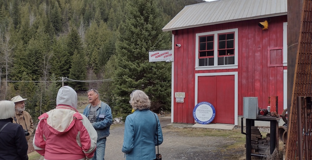A light-skinned man with a moustache, jean jacket and blue jeans speaks to four visitors in front of a wooden building painted red.