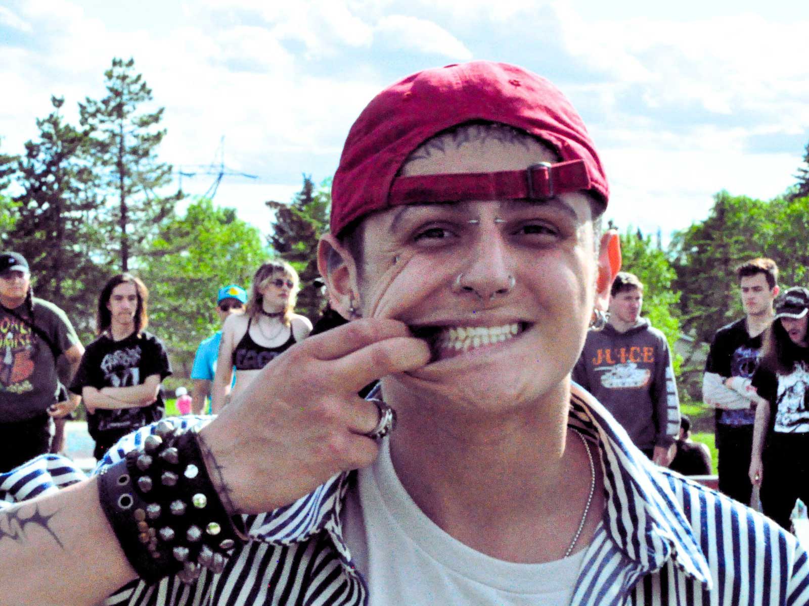 A young man wearing a backwards red ball cap puts two fingers in his mouth to pull his lips apart from his teeth, smiling. He has facial piercings and is wearing a black leather cuff with spikes. He is standing outdoors at a skatepark and several people are standing in the background.