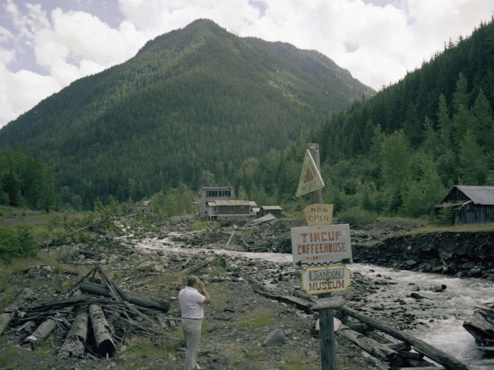 A wooden signpost directs visitors to the &ldquo;Tincup Coffeehouse&rdquo; and Sandon Museum. A few wooden buildings are visible in the distance, beyond a gravelly creek. 