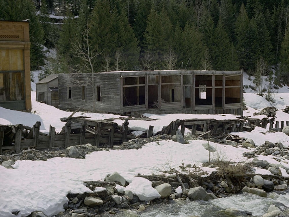 The greying remains of a former low-slung storefront sits in front of a rushing creek amid significant snow accumulation.