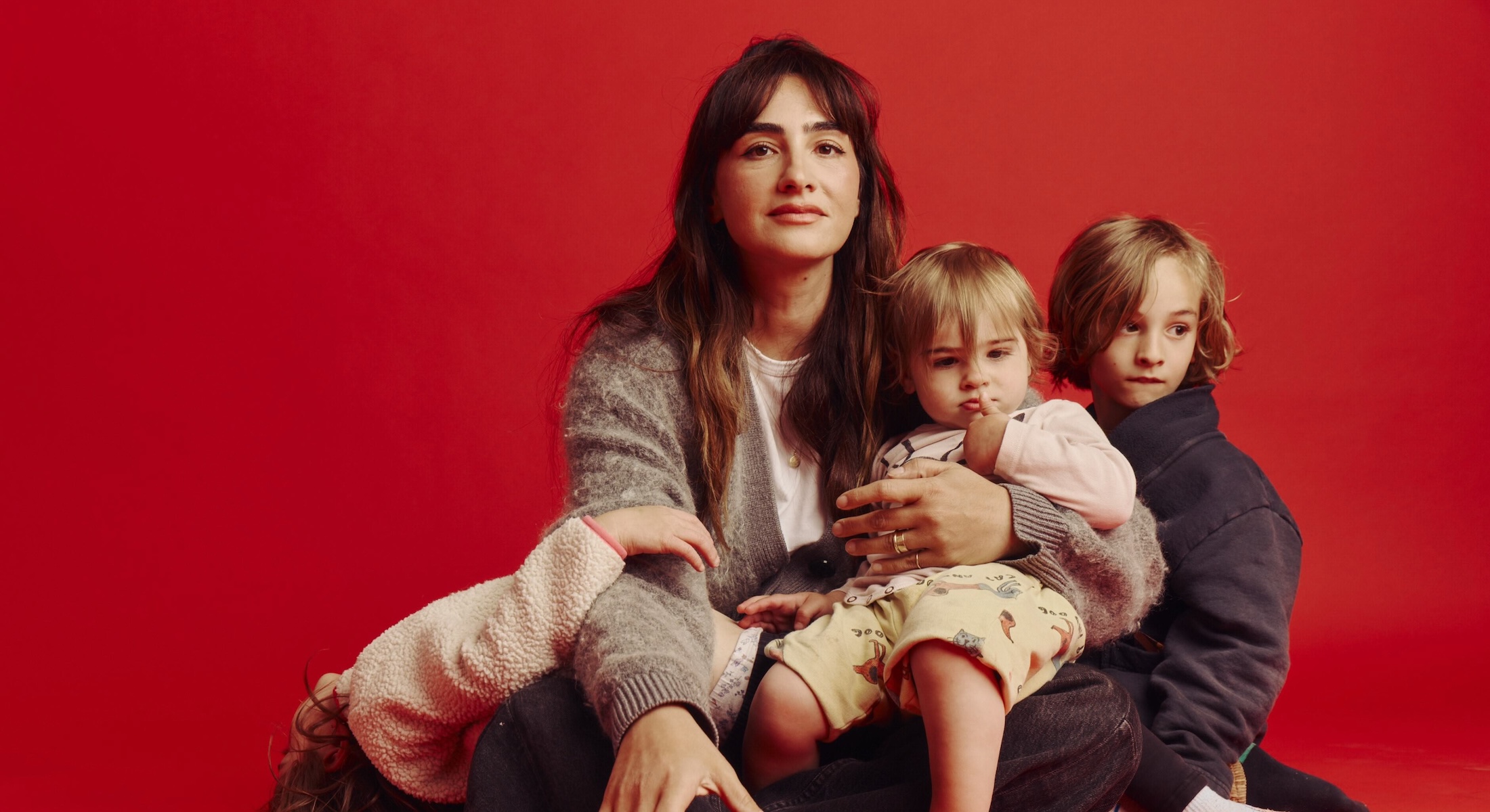 Amil Niazi is seated on the floor against a red background with her three children. She has long dark hair with bangs and she is wearing a grey cardigan over a white T-shirt and black jeans.
