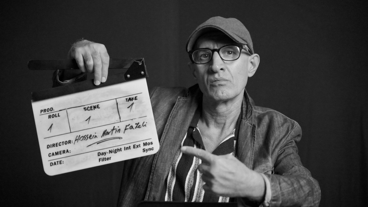 A black-and-white photo of Hossein Martin Fazeli features him holding and pointing to a director&rsquo;s clapperboard. He is wearing a ballcap, glasses and a vertical striped shirt under a dark jacket.