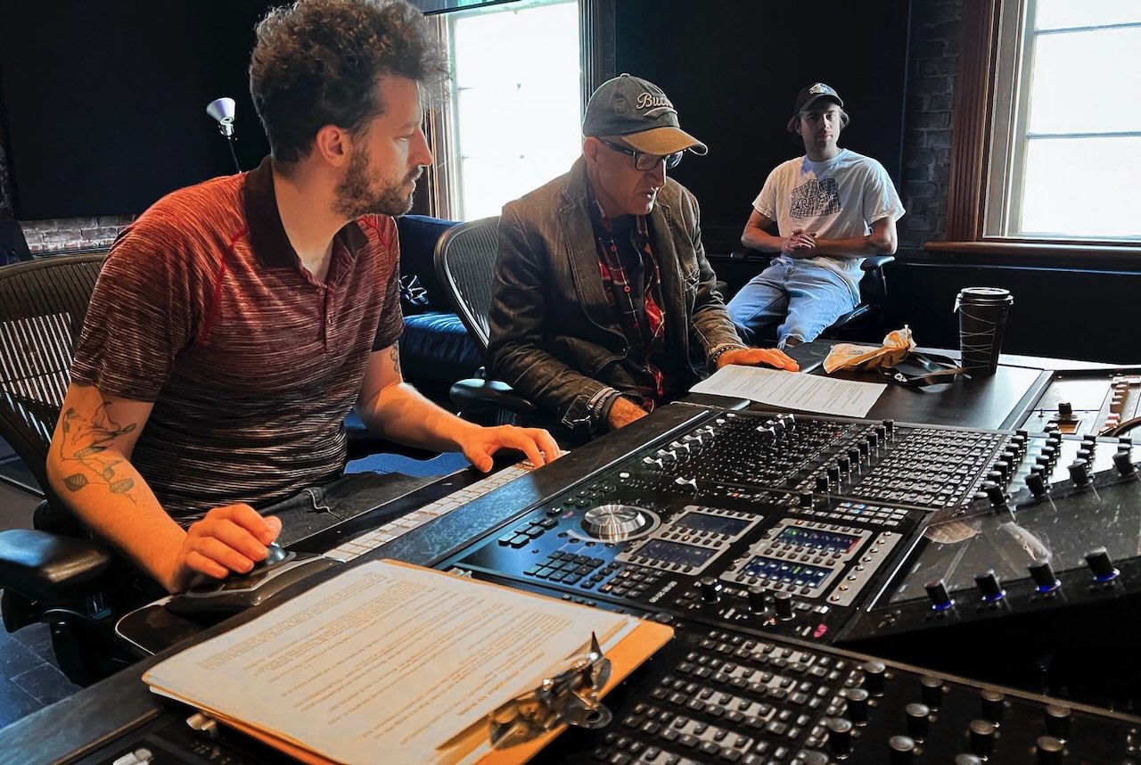 Hossein Martin Fazeli is seated at a black table outfitted with a large sound board. He is wearing a ball cap and looking down at the piece of paper on his desk. He is seated between two younger men who are part of his film crew. 