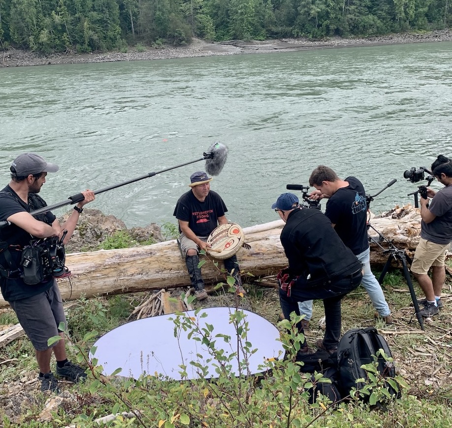 A film crew surrounds a man seated on a log holding a drum on the banks of a river. 