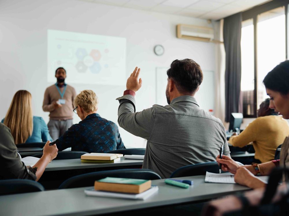 A college classroom features students seated at long grey tables with their backs turned to the camera. At the front of the room an instructor in a brown sweater stands before a white wall upon which a chart is projected.