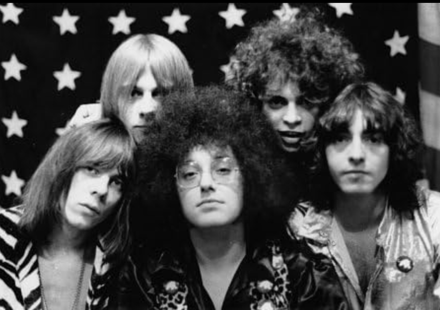 A black-and-white photograph of the members of MC5, rock musicians with long hair, posing against the backdrop of an American flag.