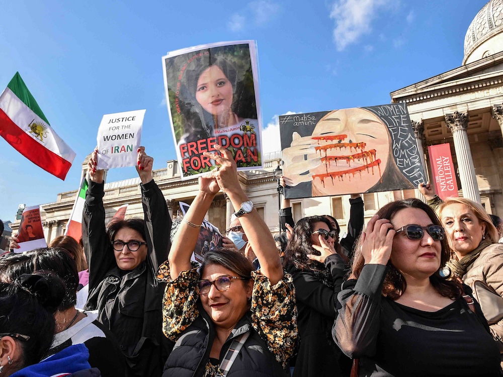 Protesters hold placards in solidarity with the Woman, Life, Freedom movement in Iran at an outdoor demonstration. They are standing close together in front the National Gallery, London, an ornate white building. The sky behind them is blue.