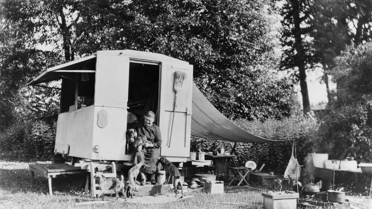 A 1934 black-and-white photo features Emily Carr seated on the front steps of her caravan in the woods. She holds a monkey in her lap and four dogs surround her.