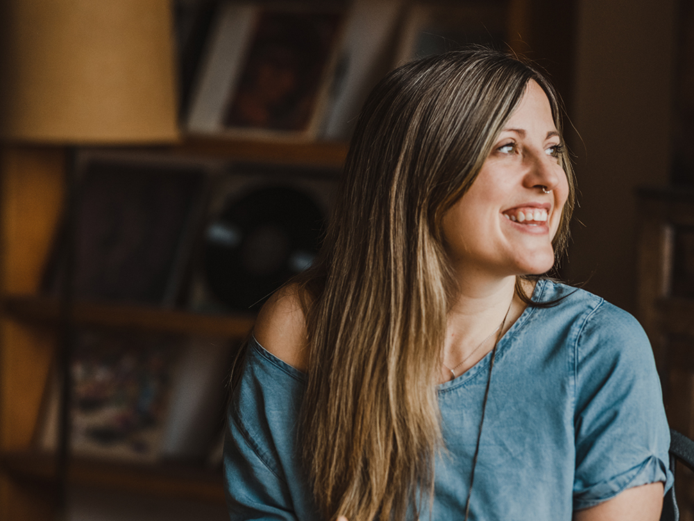 Alina Cerminara has long blond hair and is looking to the right of the frame, smiling. She is seated indoors in front of a shelf containing books and records.