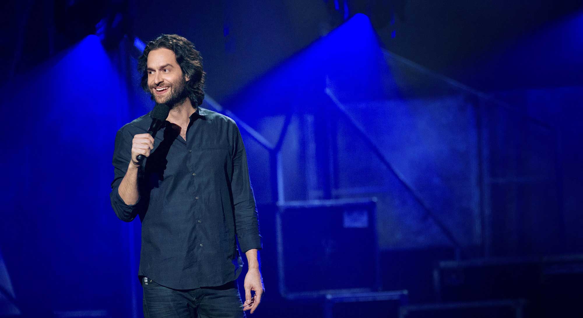 Chris D&rsquo;Elia stands to the left of the frame, smiling. He is holding a wireless microphone and standing onstage with blue lights in the background. He has dark wavy hair and a beard. He wears a dark button-down shirt and black jeans.