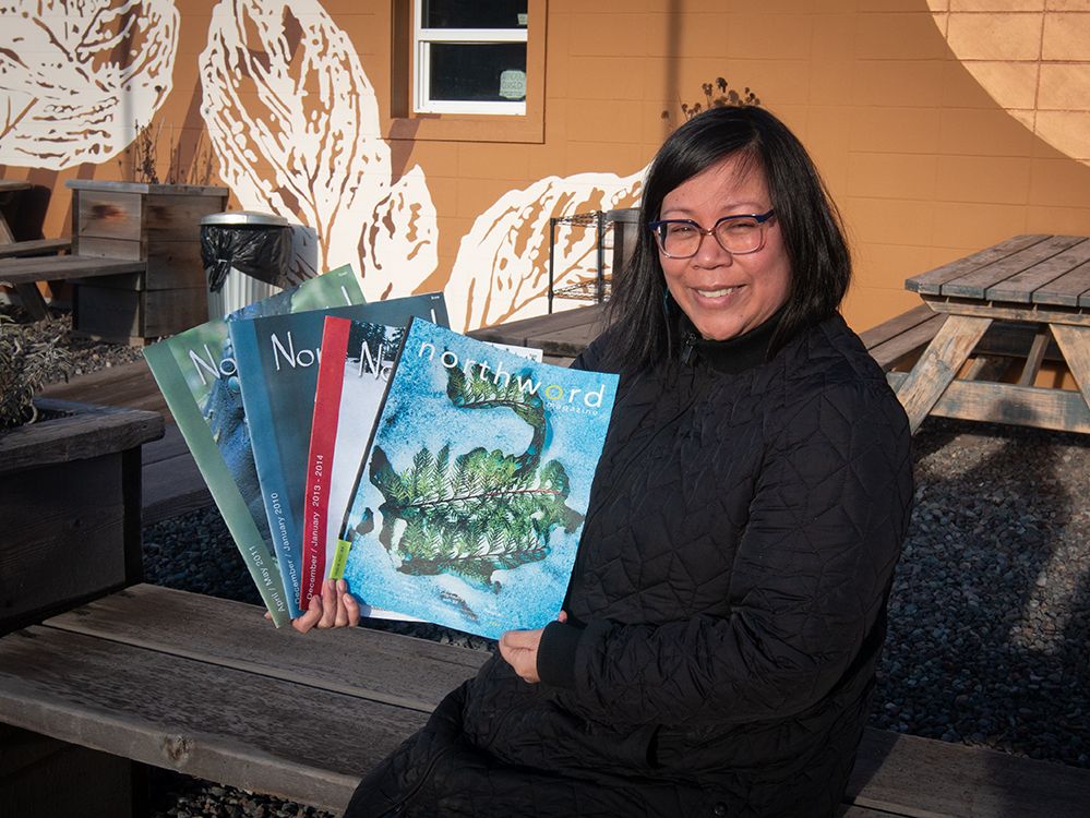 Christine Anonuevo has dark shoulder-length hair and glasses. She is smiling and holding several copies of Northword magazine. She is seated on a wooden bench outside a caf&eacute;.