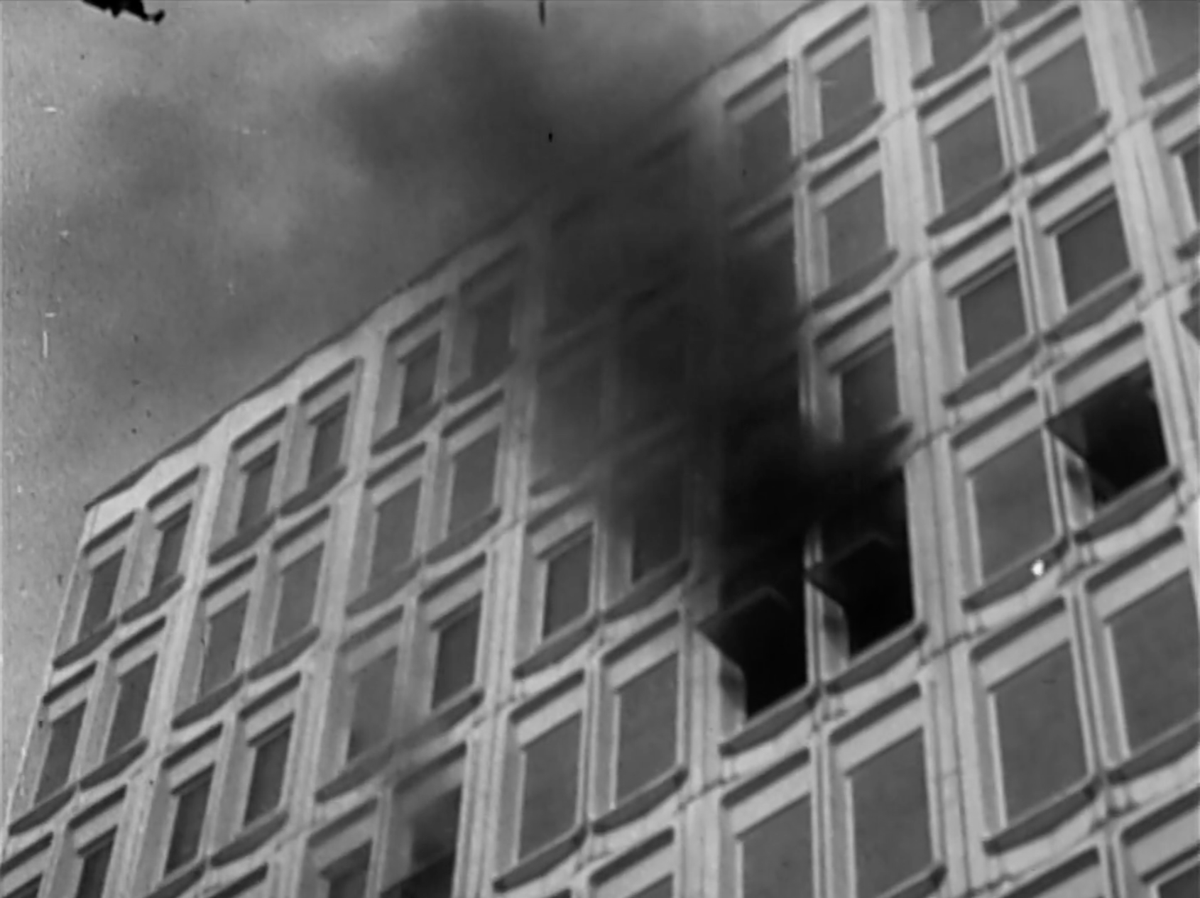A black-and-white archival photo of a tall building with rows of windows has smoke billowing out of two of them.