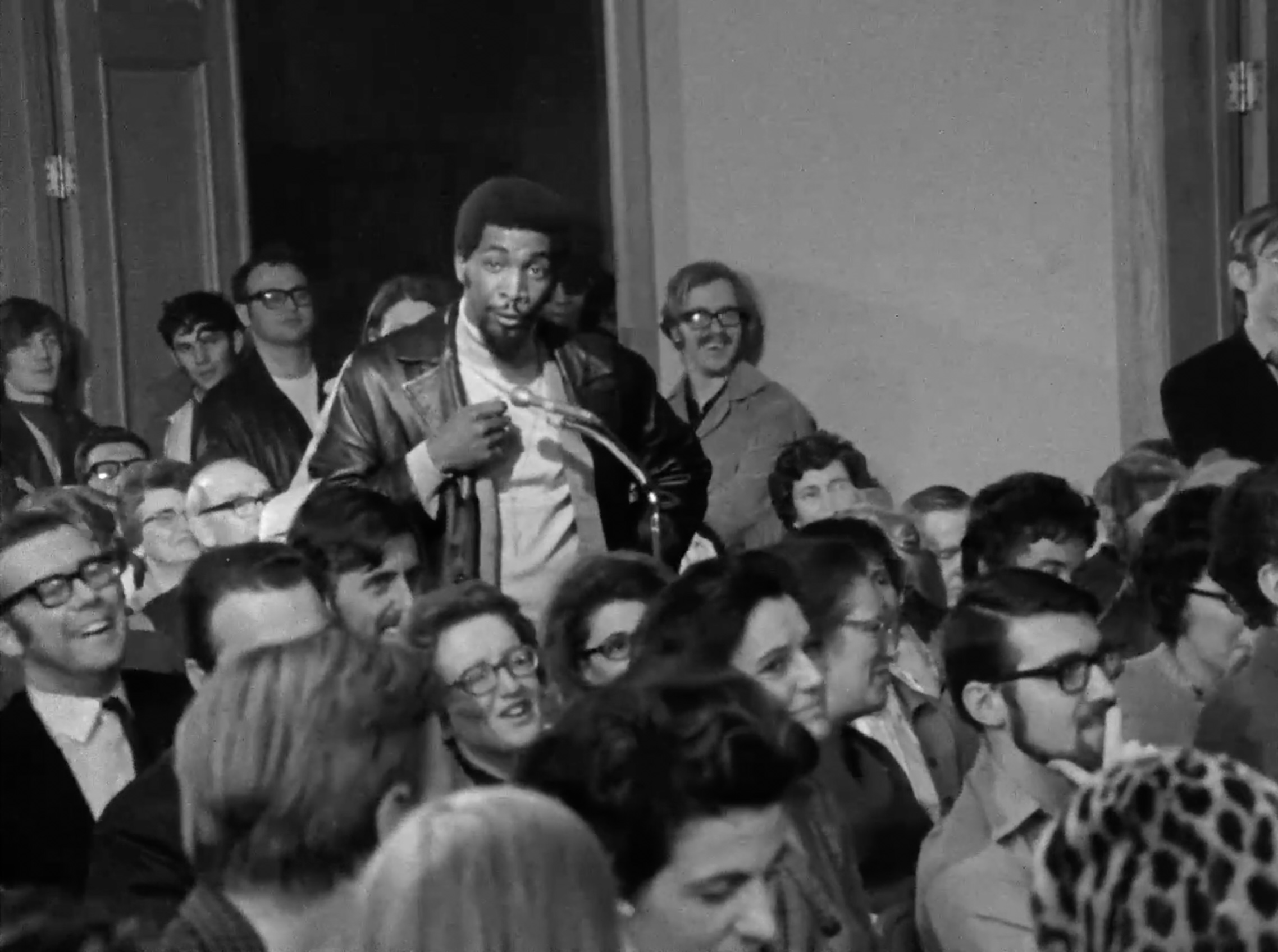 A young man stands in a crowded room to speak. He is standing, mid-speech, at a microphone while others are seated around him. He is the only Black person in the frame.