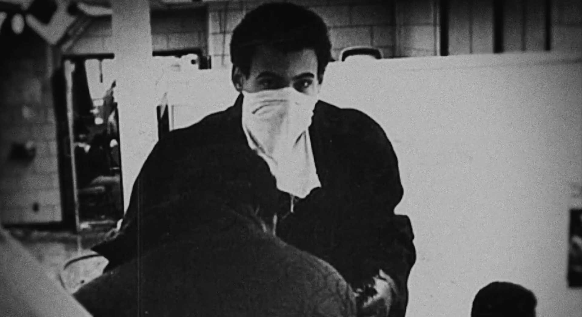 A young man in a dark winter coat wears a white bandana covering his face. He looks at the camera while a person stoops in front of him.