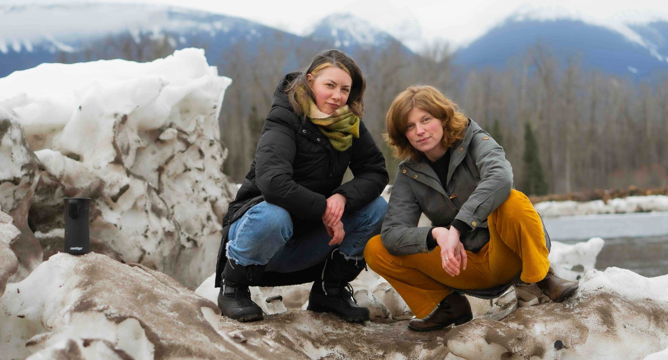 Pamela Haasen, left, and Naomi Kavka pose together outdoors, crouched on a pile of old snow. Behind them is a stand of trees and a blue mountain range. Haasen is wearing a black winter jacket and blue jeans; Kavka is wearing a grey winter jacket and yellow trousers.