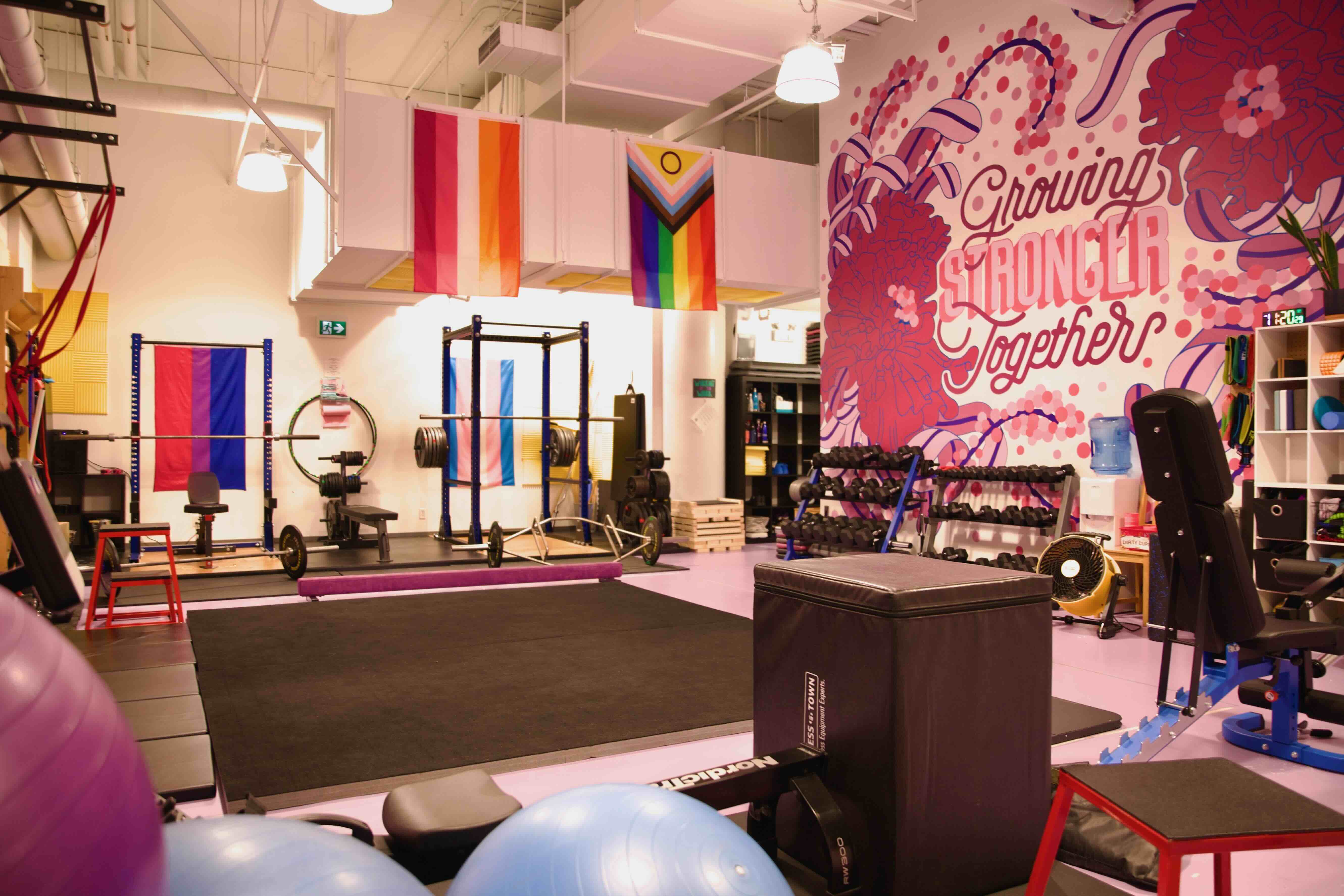 The interior of the Witness the Fitness gym features a black mat at the centre of the floor and several types of fitness equipment. Pride flags hang on the walls and a pink mural that says &ldquo;growing stronger together&rdquo; is on the wall to the right of the frame.