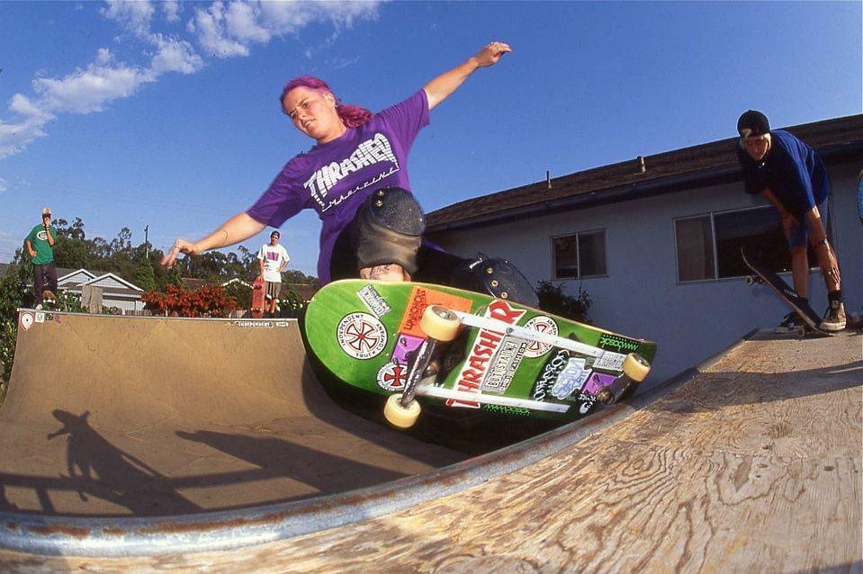 JoAnn Gillespie is riding her skateboard on a wooden mini-ramp. She is a white woman in her twenties with purple dyed hair in a ponytail, wearing a purple t-shirt that says Thrasher. She wears black shorts and black knee pads for protection. Her skateboard is green, covered in skateboard stickers. There are three young men in the background watching her perform a grind on the ramp.