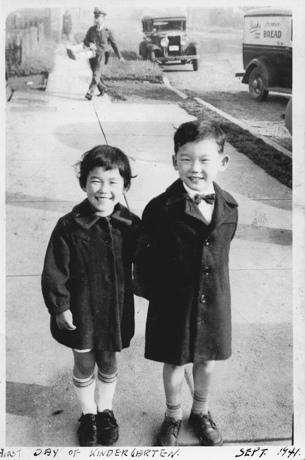 A black and white photograph of two five-year-old children in 1941. On the left, Marcia Suzuki has dark hair pulled back with bangs. She is wearing a dark jacket and white knee socks. She is standing next to her twin brother David, at right, who is taller than her. He is wearing a dark jacket and shorts.