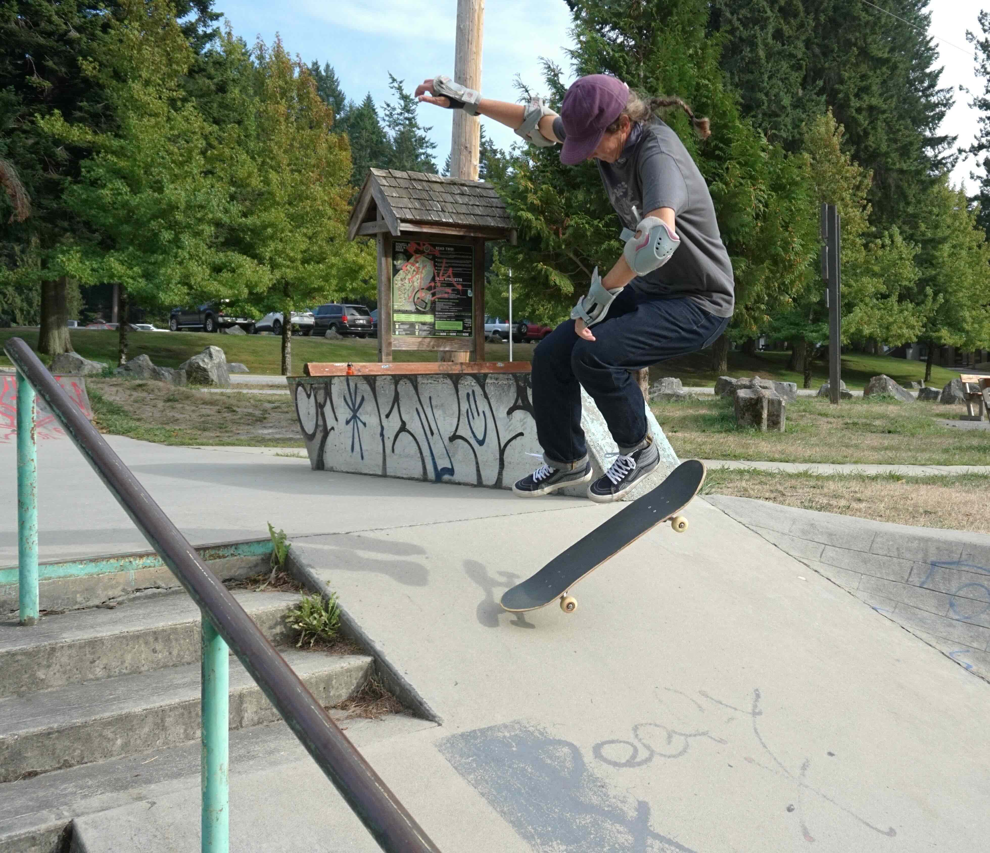 A colour photograph of Natalie Porter, a middle-aged white woman with brown hair in a braid. She is wearing a purple hat, gray t-shirt and blue jeans, and has elbow pads and wrist guards for protection. She is skateboarding on a concrete bank with trees in the background.
