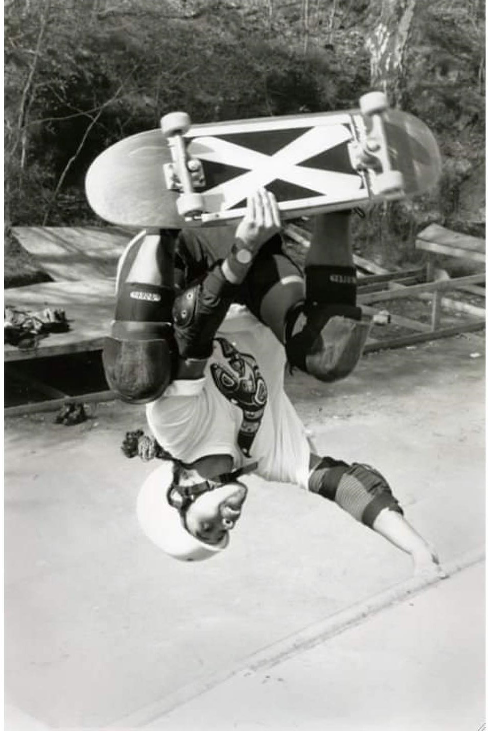 A black and white photograph depicts Stephanie Person riding a skateboard in a vert ramp performing a handplant. Person wears protective gear including a helmet, knee pads and elbow pads. She has a dark skin tone and is wearing a white t-shirt with an Indigenous motif of a whale and black shorts.
