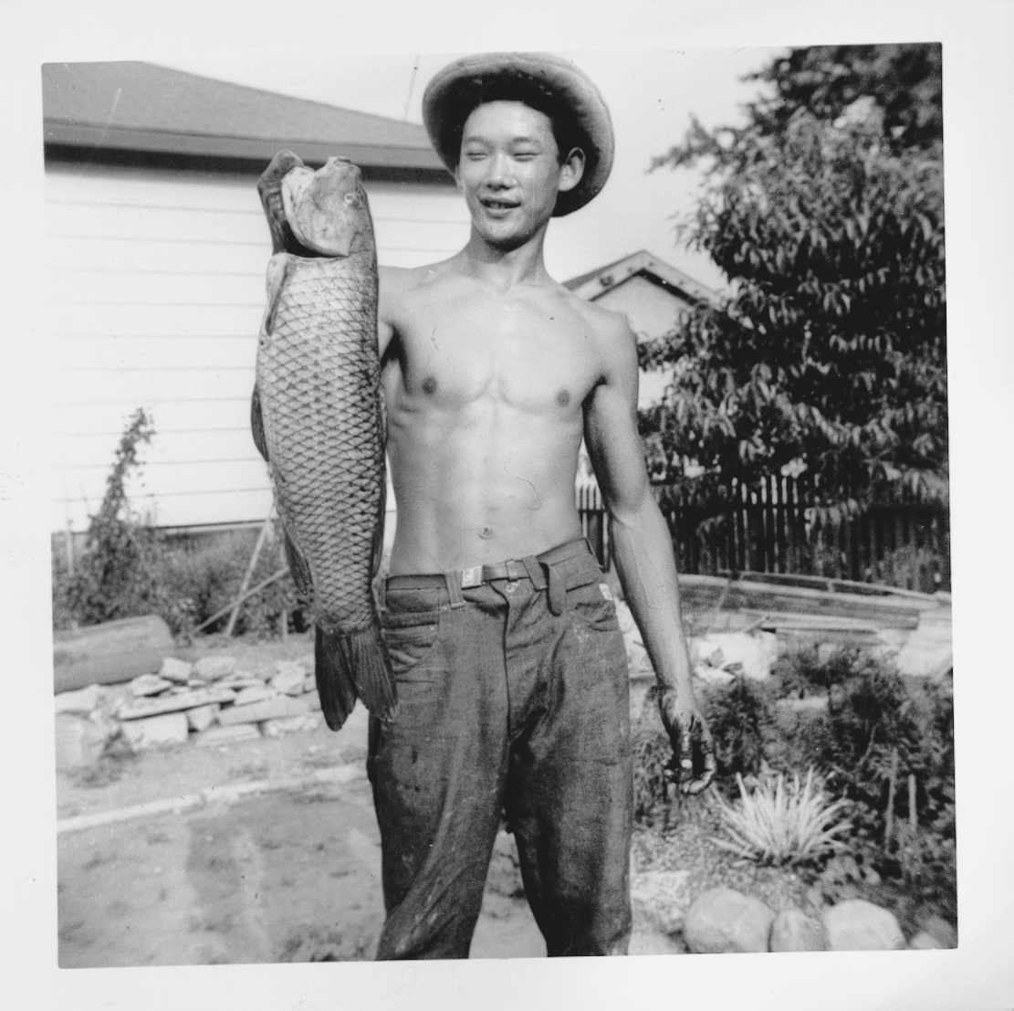 David Suzuki is a young man wearing a summer hat and trousers with no shirt. He is holding a large fish and standing in the yard of a house.