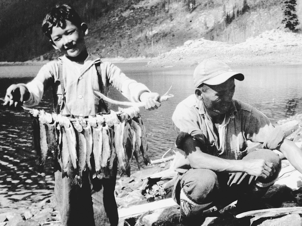 A black and white photograph of David Suzuki as a young boy on the shore of a lake. He stands to the left of the frame wearing suspenders and a button-down shirt. He holds a large bounty of fish strung on a line. Next to him, at right, is an older man in a ball cap, crouching and working on an item out of frame.