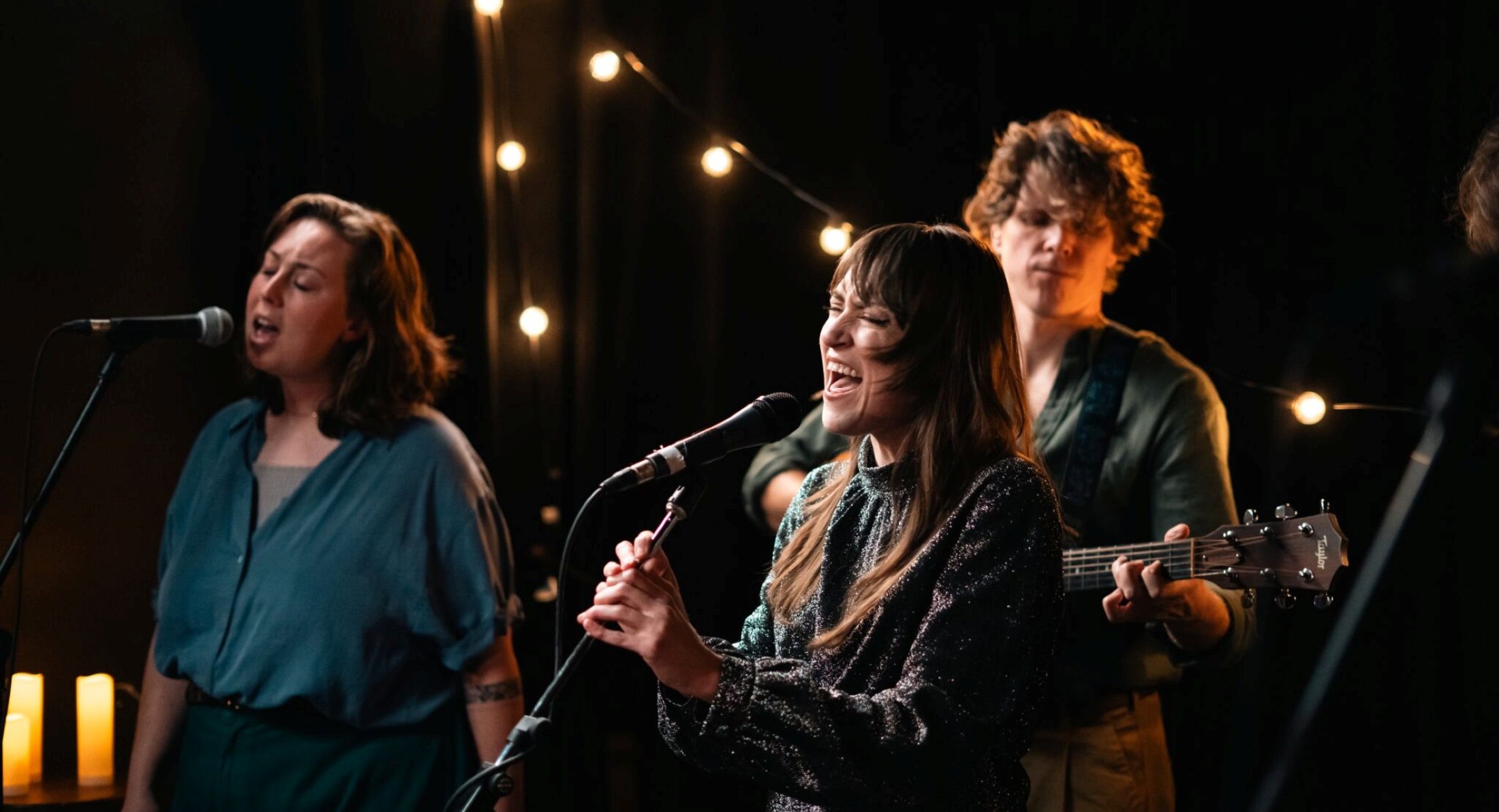 Cassidy Waring, centre, sings into a microphone. She has long brown hair with bangs. She is standing onstage beside another vocalist and in front of a guitar player.