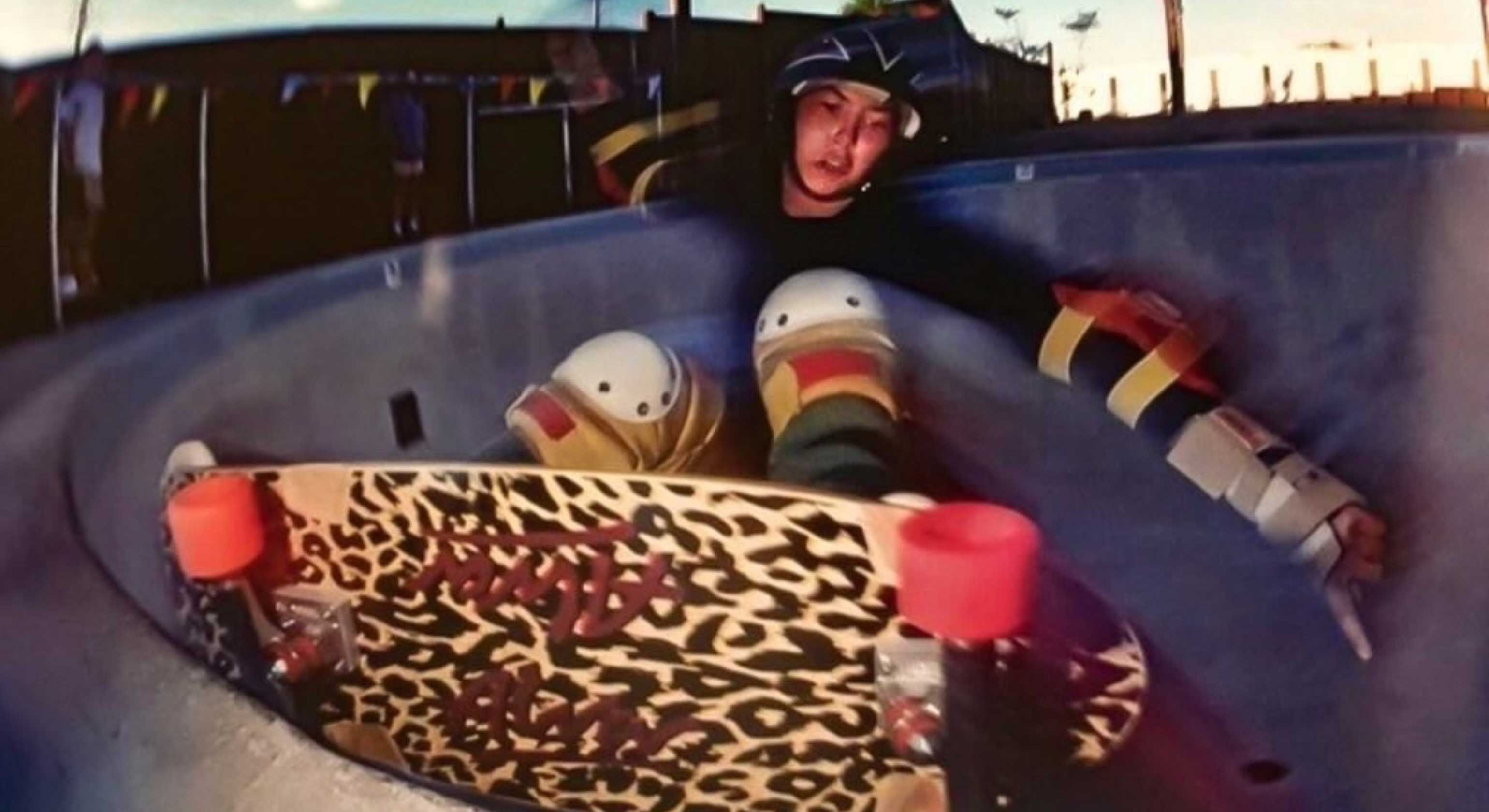 A colour photograph using a fisheye lens features a skateboarder skating at dusk while the sun leaves the sky behind them. Terry Lawrence is wearing a helmet and protective equipment on their arms and legs while they skateboard against the edge of the bowl of a skatepark. The bottom of their skateboard faces the camera, revealing a leopard print pattern.