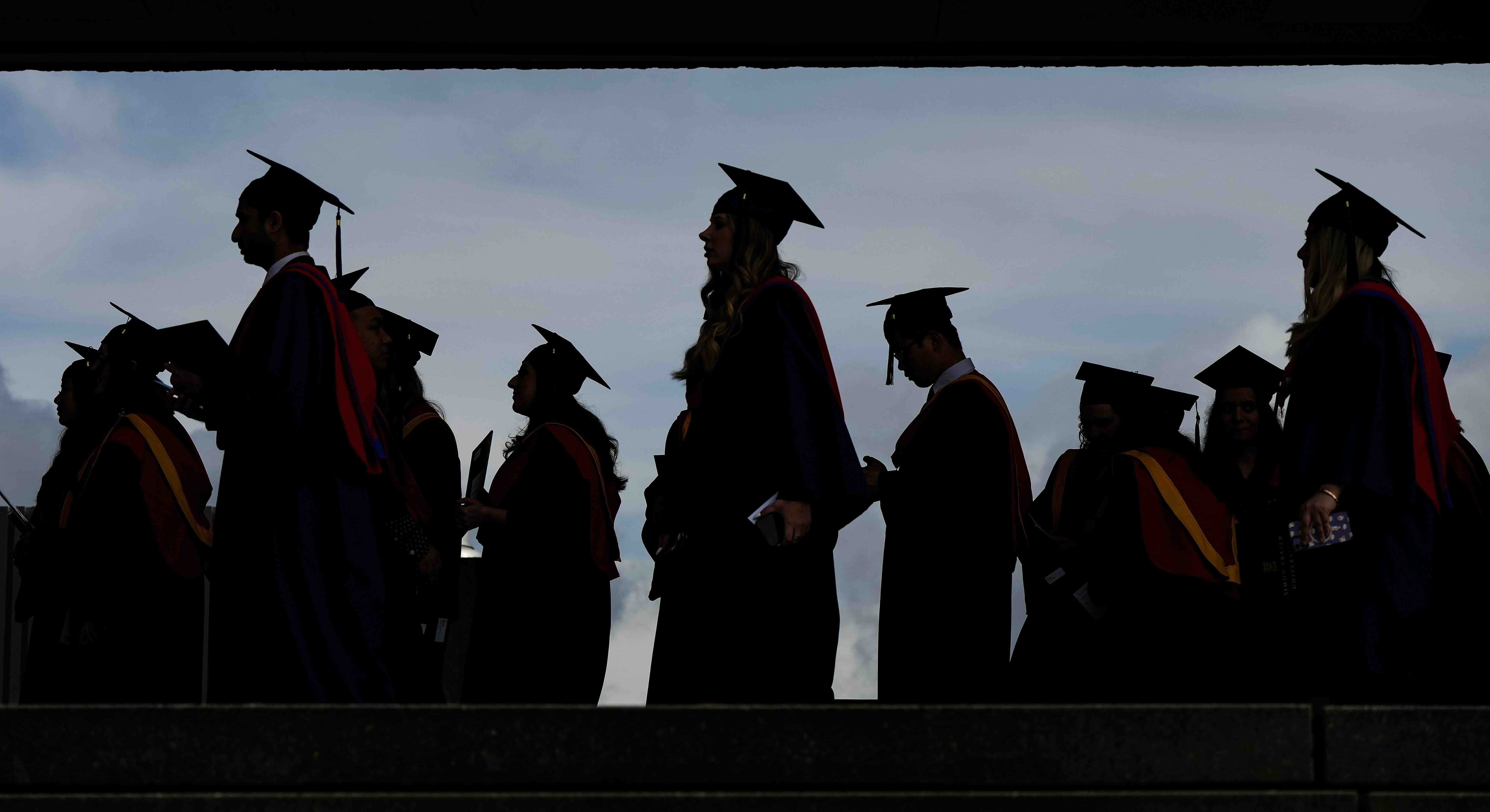 Graduates in caps and gowns stand in silhouette against a grey sky. They are standing below a thick concrete ceiling shown in silhouette.