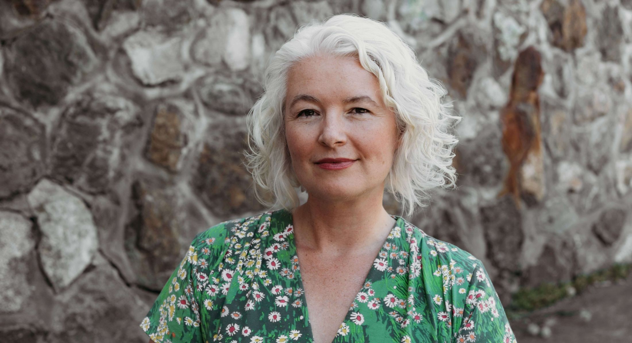 A middle-aged white woman with chin-length curly silver hair looks directly at the viewer. She wears a short-sleeved green floral dress. There is a blurred stone wall behind her.