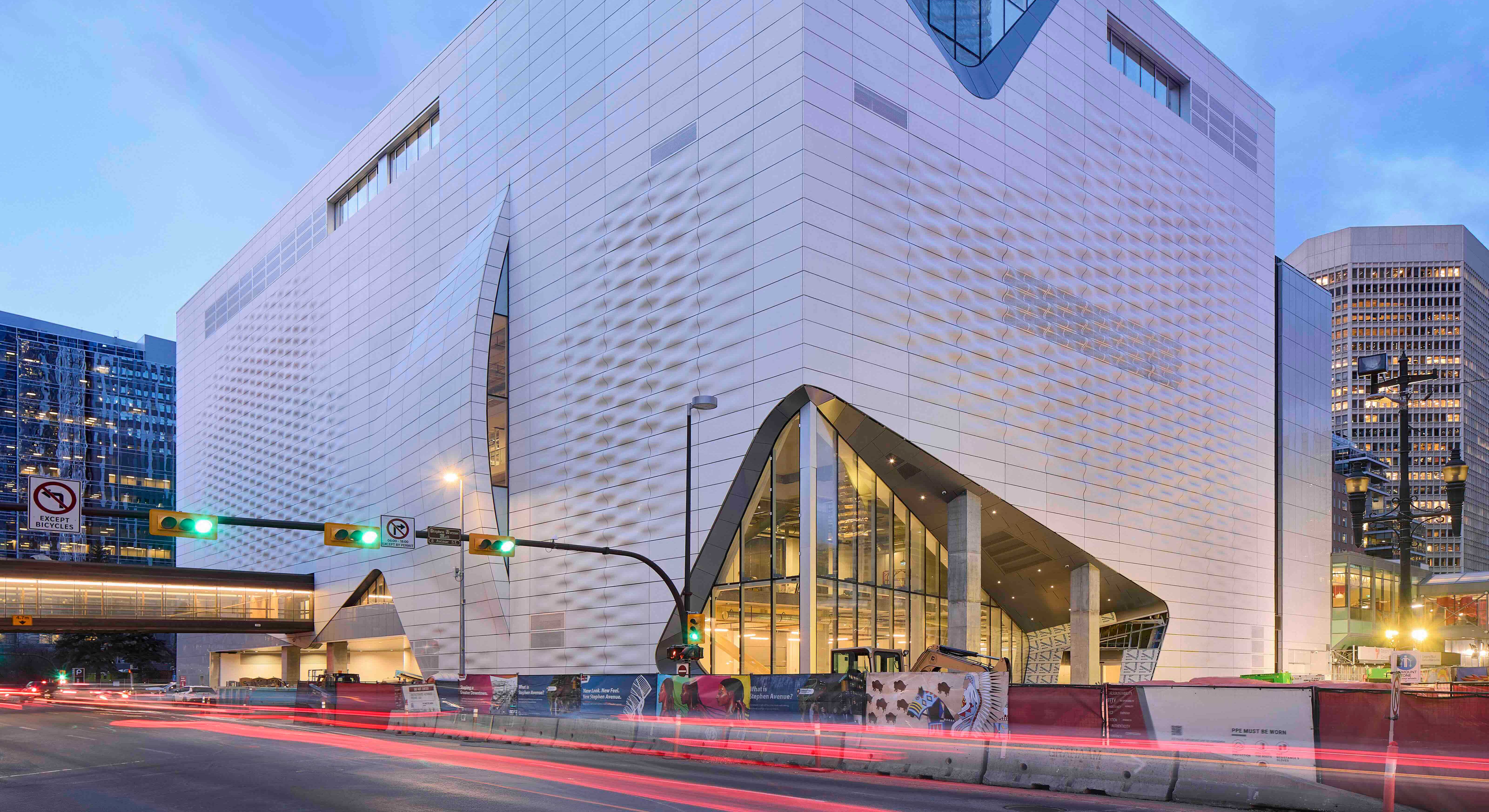 A large white building with contemporary textured walls stands on the intersection of a downtown street at dusk. Blurred red lights of cars are on the street below. 