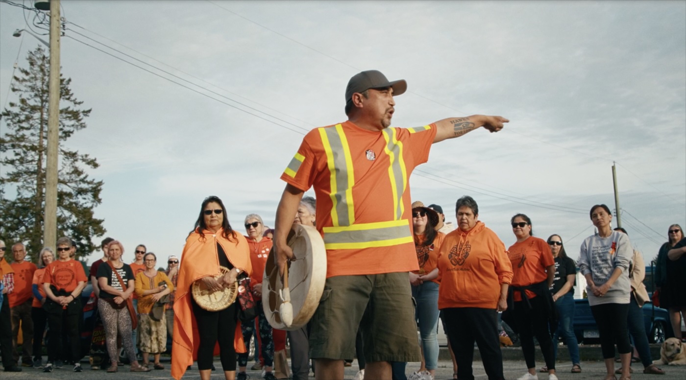 Scott Galligos wears an orange safety shirt and holds a drum. He is pointing at a crowd of Tla&rsquo;amin Nation members and residents of the City of Powell River, most of whom are wearing orange shirts. 
