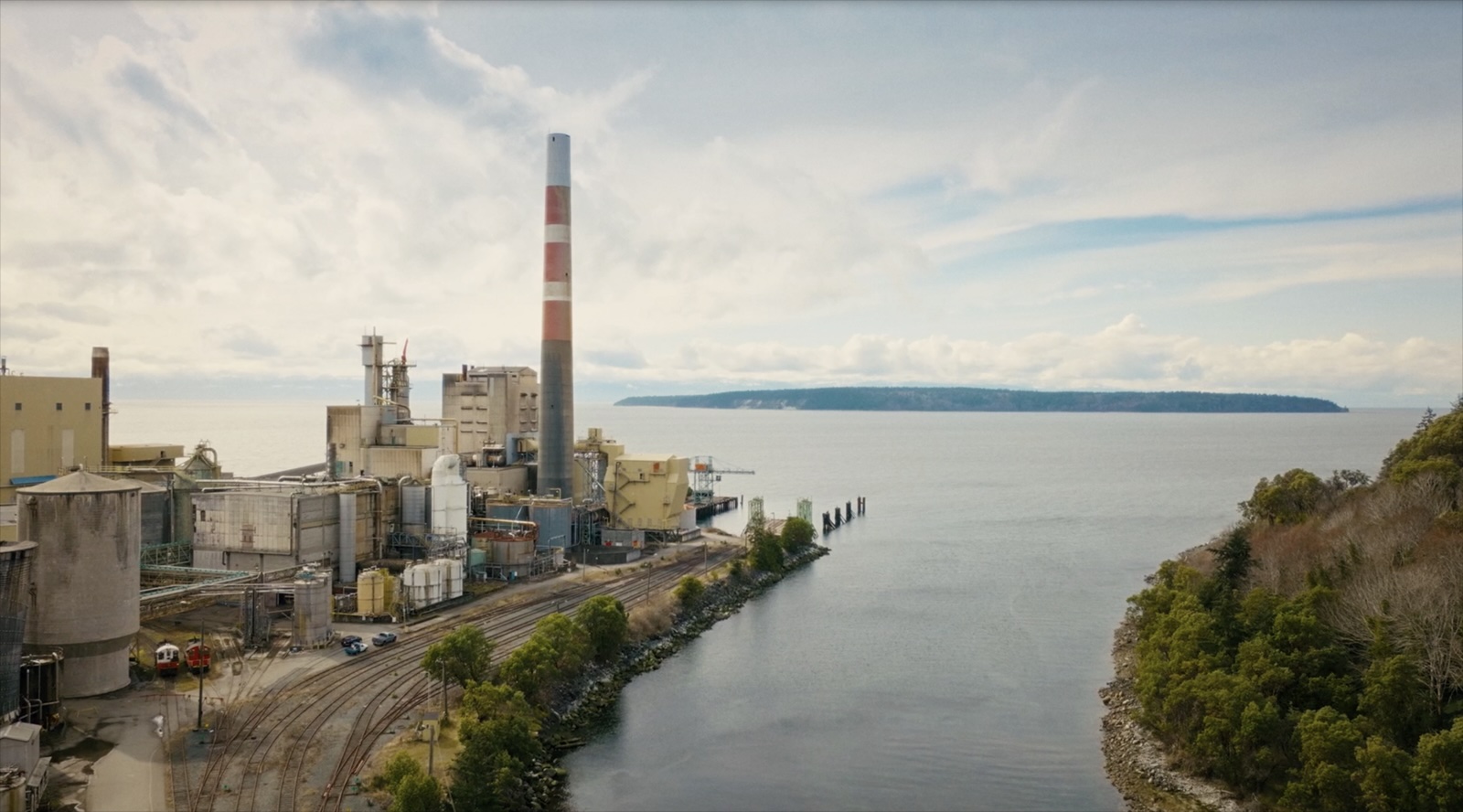 A mill smokestack, buildings, and old railway track to the left; a big flat river in the centre; a treed bank to the right.  