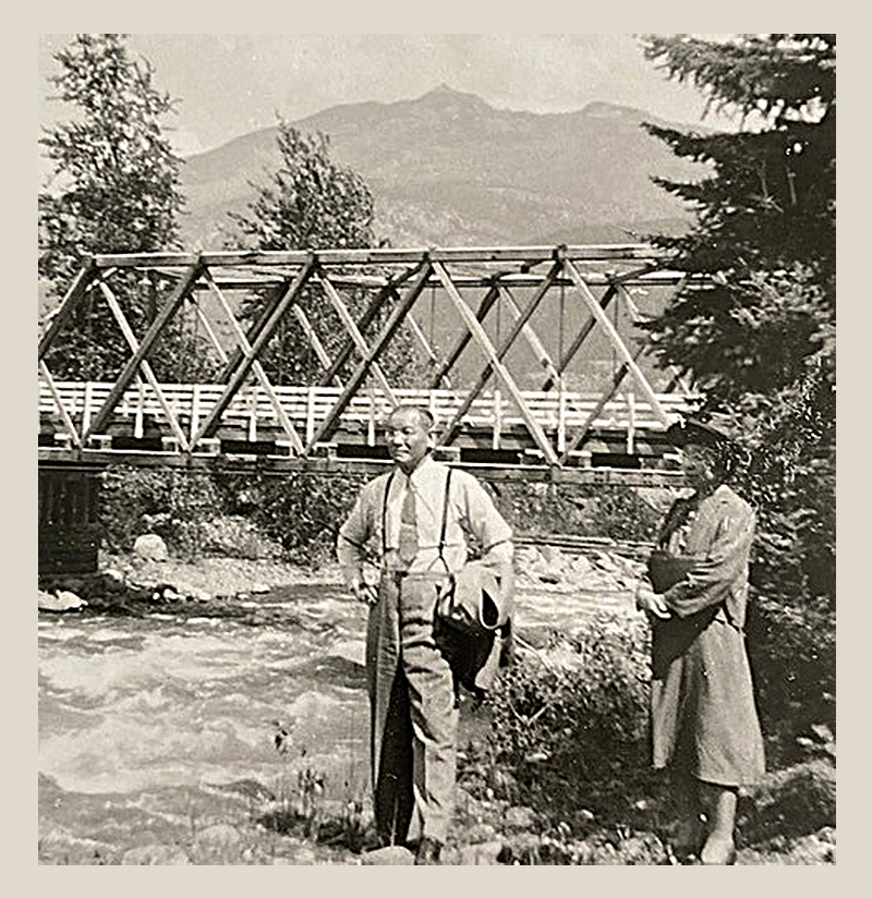 A sepia-toned historical photo of Bunjiro and Kimi Uyeda, standing by a fast-moving river with a trestle bridge behind them.