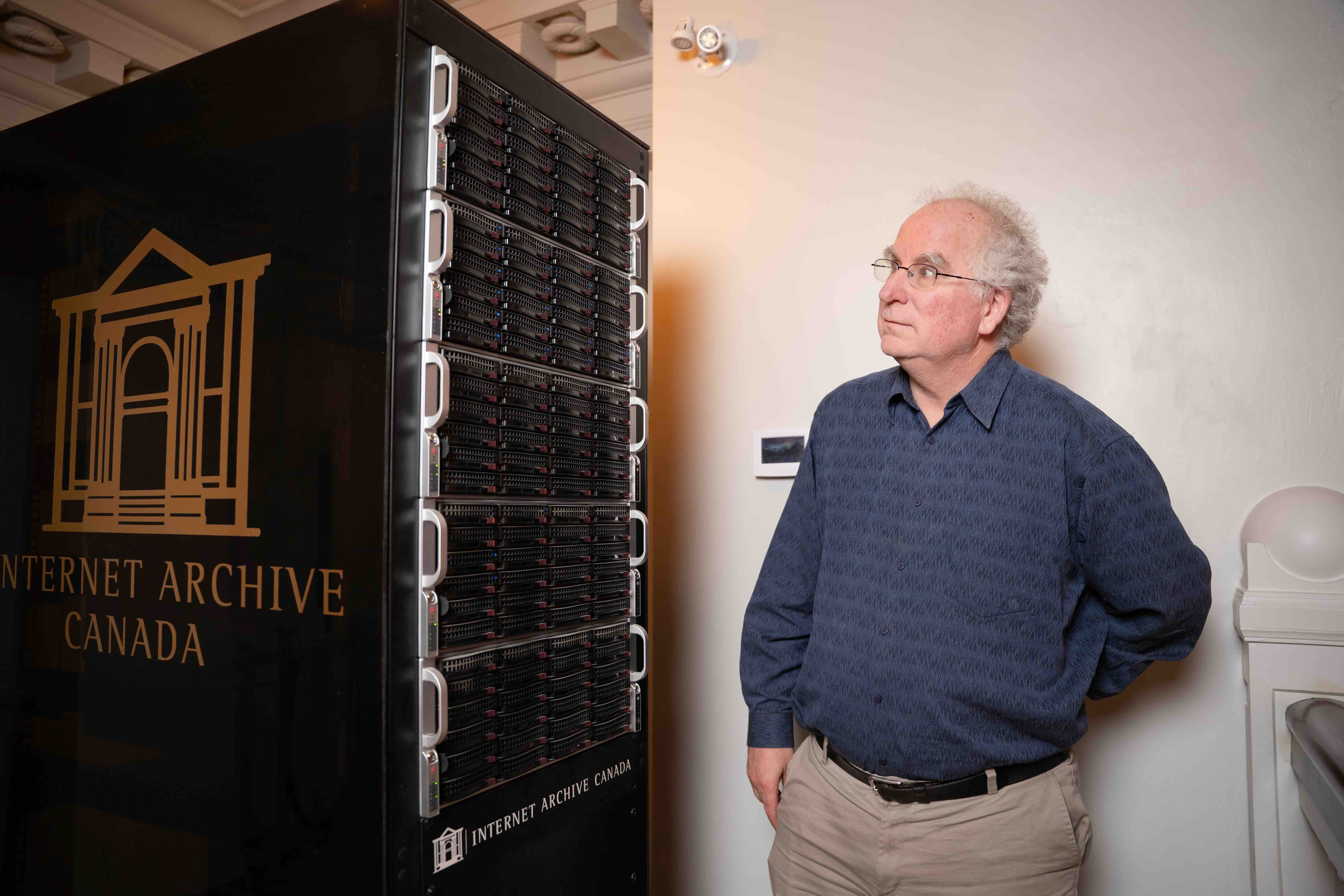Brewster Kahle is standing to the right of the frame in an indoor space against a white wall. He has short curly white hair, glasses, a blue button-down shirt and khaki coloured trousers. To his left is a large rectangular black unit with &ldquo;Internet Archive Canada&rdquo; inscribed on it. They are some of his Canadian servers for Internet Archive Canada.