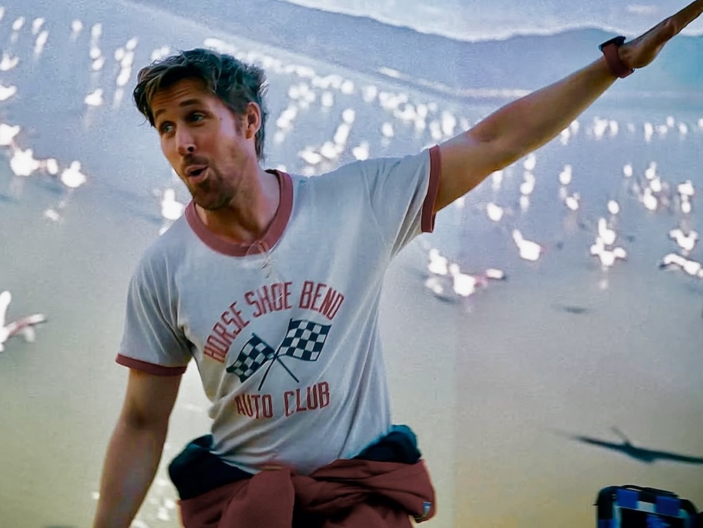 Actor Ryan Gosling has short blond hair and light facial hair. He is in a dimly lit room, spreading his arms wide, like an airplane, and standing against a wide projected background of objects on a beach.
