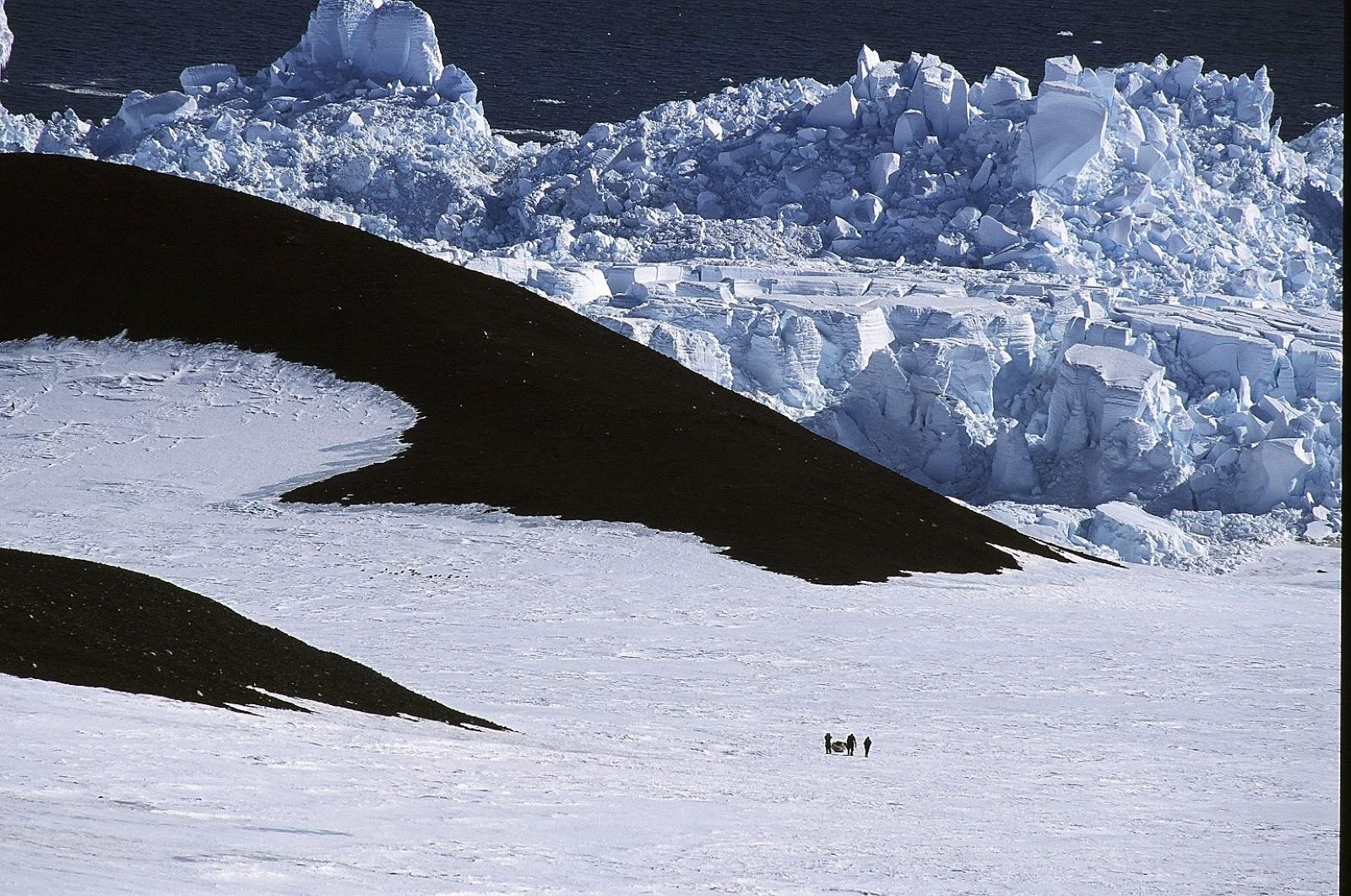 A panoramic photograph depicts vast black rocky outcroppings from fields of ice and snow; the ocean is in the background towards the top of the frame. In the lower right of the frame, three human figures are visible in silhouette, walking and pulling a sledge of supplies.