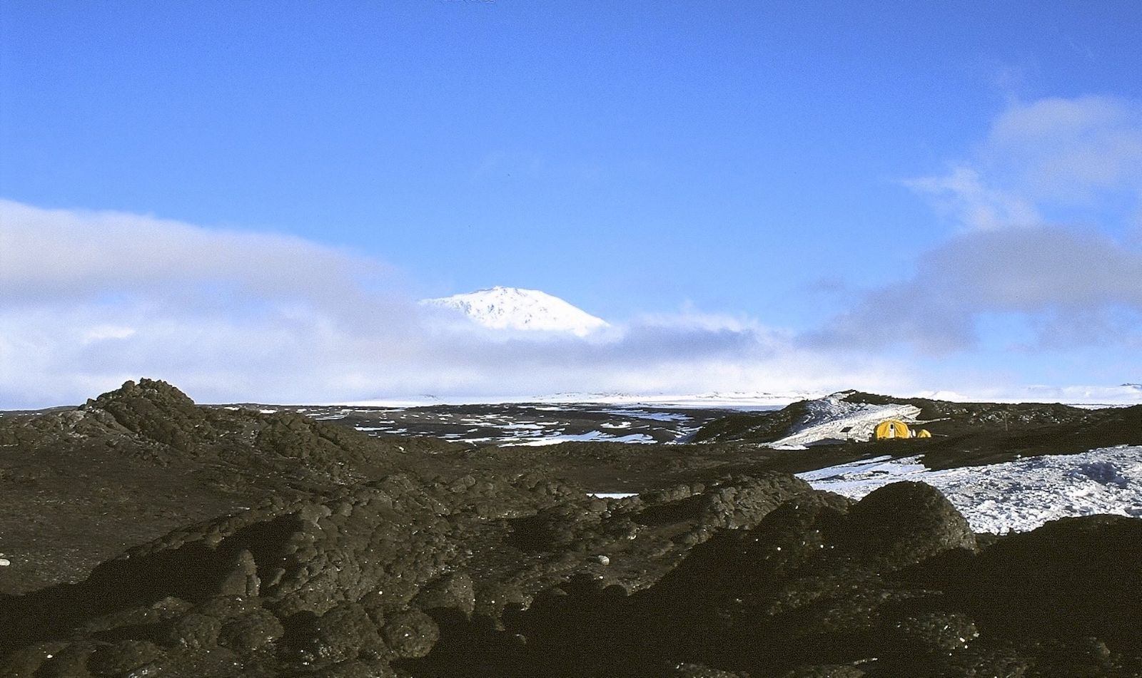 A panorama photograph of a dark rocky landscape shows a small collection of rounded yellow tents to the right of the frame. The tents are surrounded by patches of snow and ice. There is a white snow-covered mountain in the background; a band of clouds moves across its base against a bright blue sky.