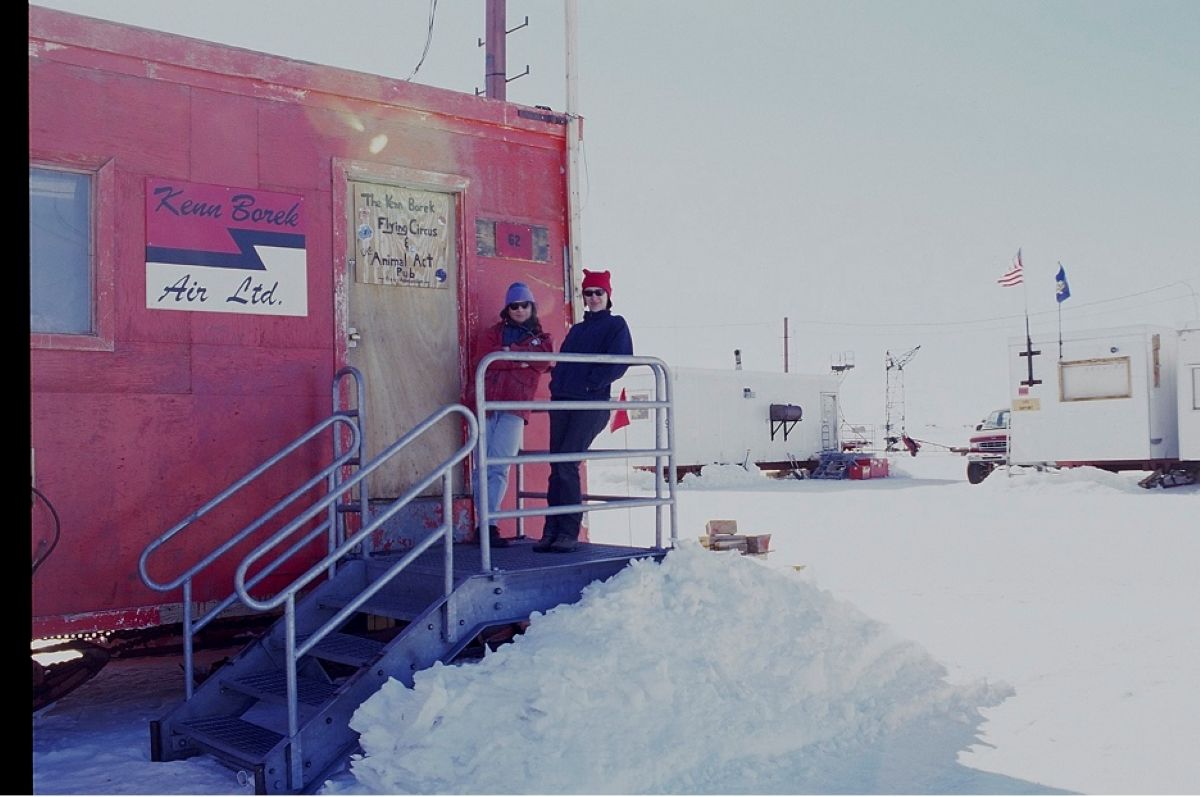 Two women stand at the top of a short set of grey metal steps in front of a light brown door of a red trailer. A sign to the left of the frame on the trailer reads &ldquo;Kenn Borek Air Ltd.&rdquo; The women are wearing sunglasses, toques and casual winter clothing.