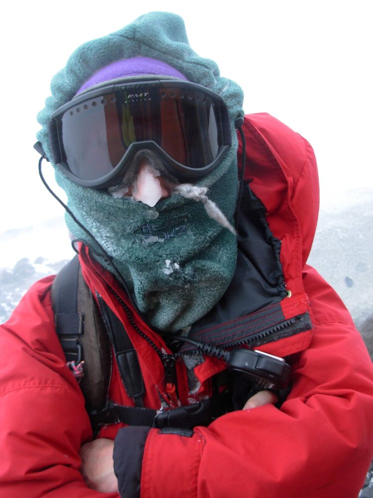 Louise K. Blight is wearing ski googles, a green fleece hood and a backpack with a bright red parka. Her nose is visible, and covered in snow. Behind her is a dark rocky landscape and a white sky.