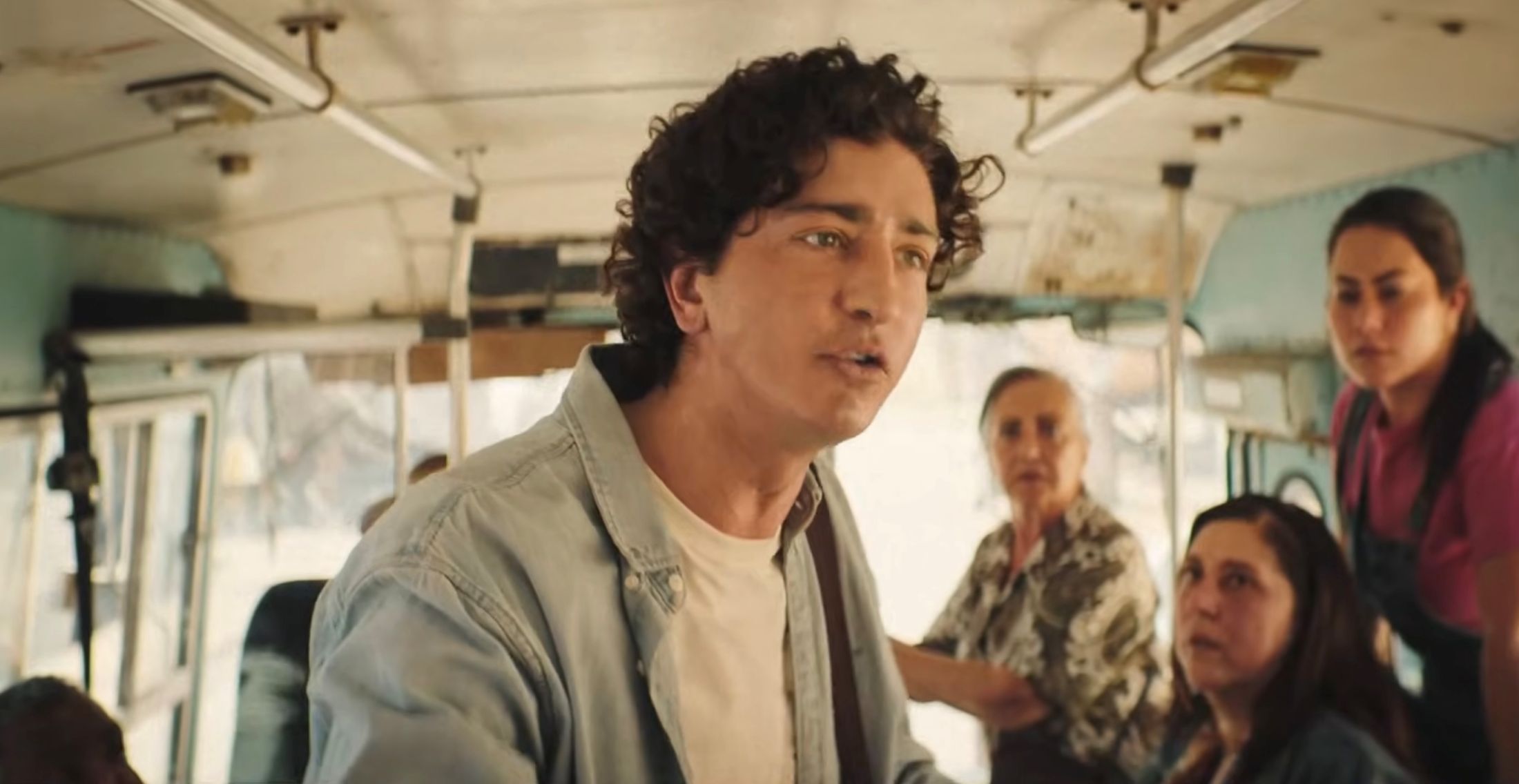 Actor Johnny Massaro has dark curly brown hair and he is wearing a blue button-down shirt over a white T-shirt. He is standing inside a public bus speaking urgently to passengers.