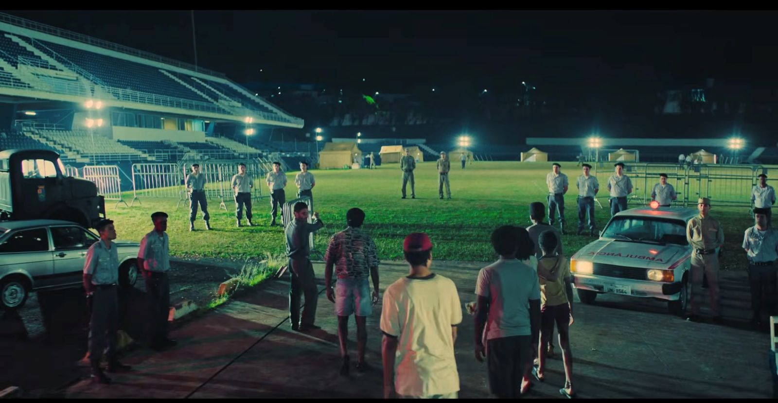 Men in military uniforms and berets stand in the grass of a soccer stadium as people arrive by ambulance at night.