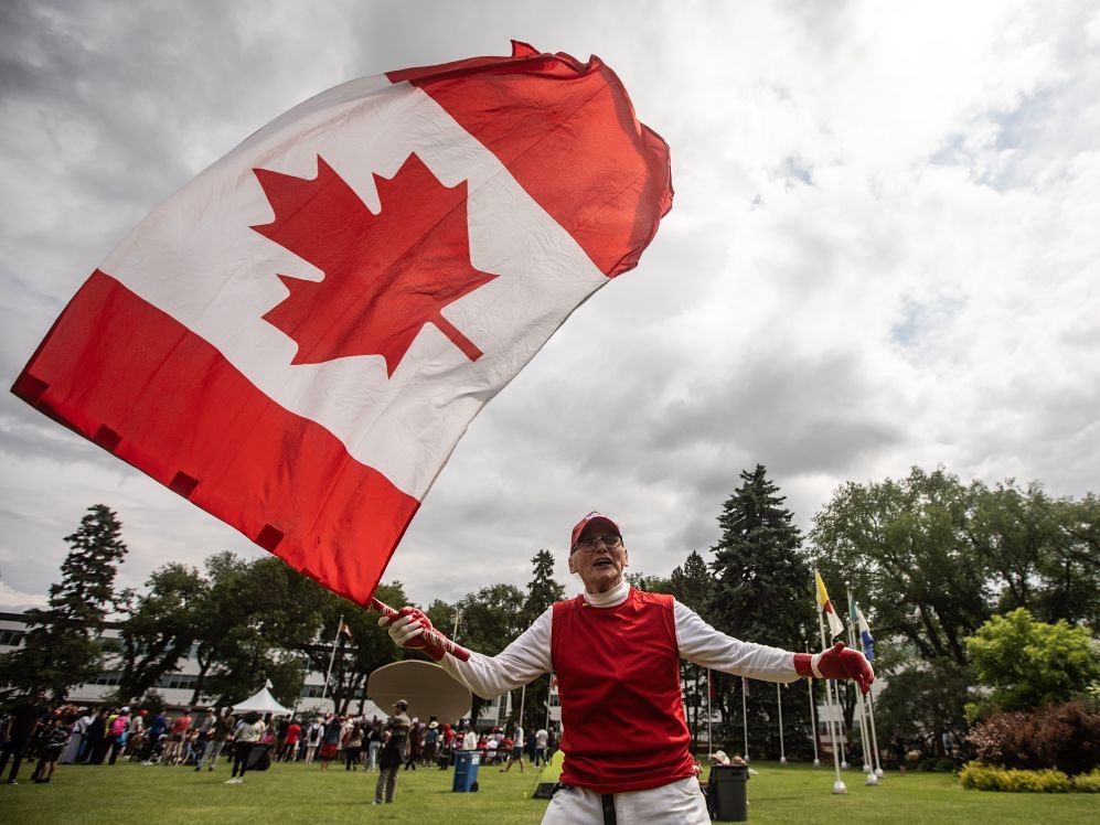 A person wearing red and white waves a large Canadian flag at an outdoor Canada Day celebration.
