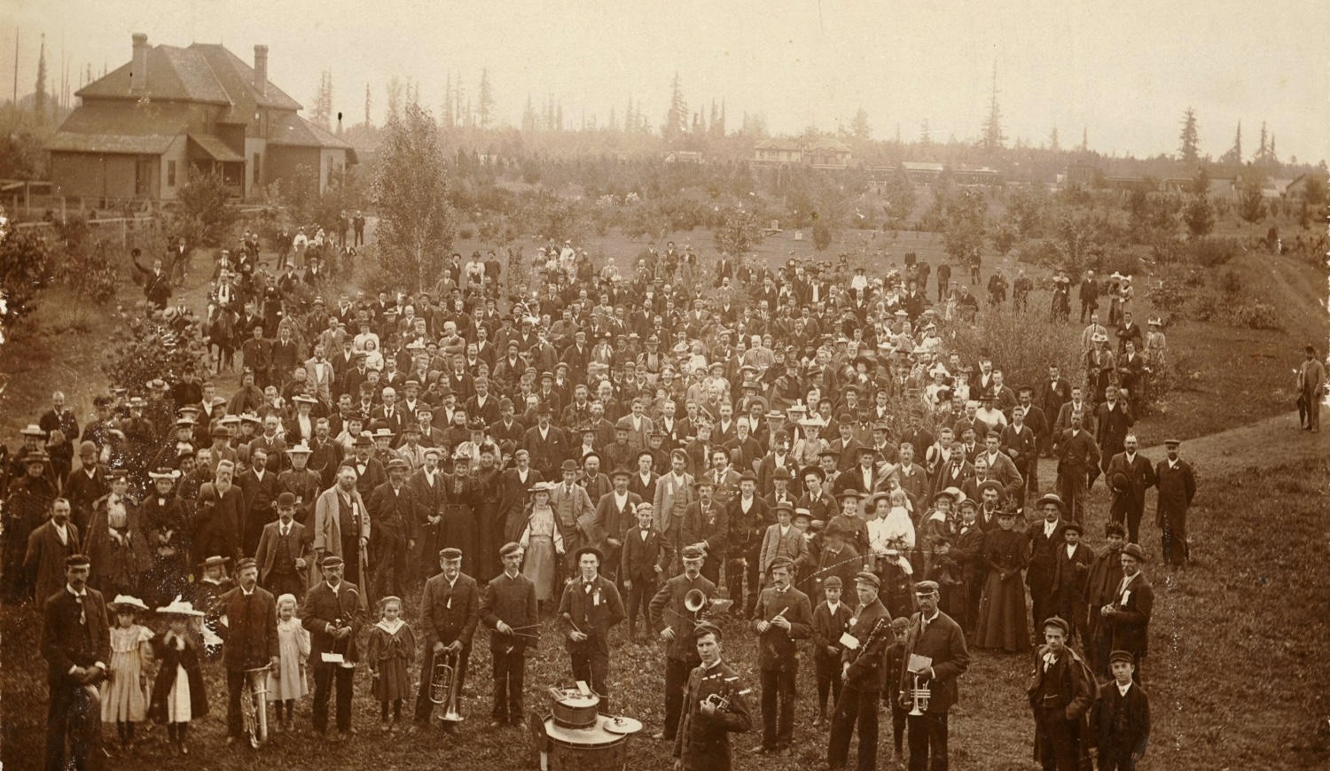 An old sepia-toned photo of a large crowd of people, many clad in business attire, posing for a group photo with a structure in the background. A brass band stands at the front of the picture.