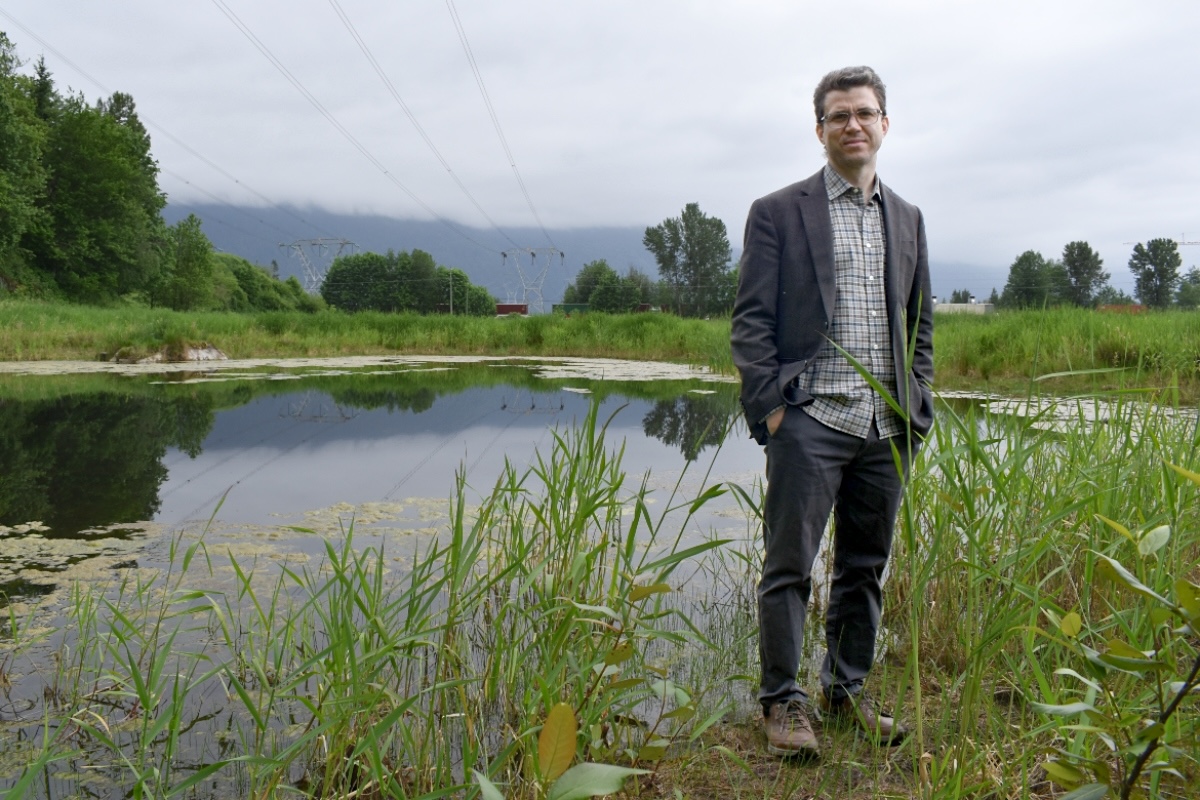 A man with light skin wearing a blazer over a checkered shirt and long pants stands next to a small pond surrounded by grasses.
