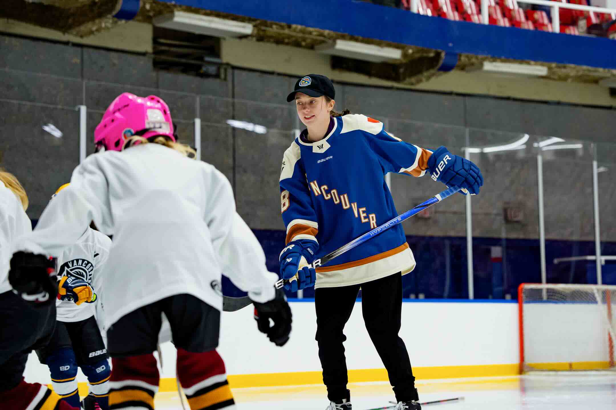 A female hockey player skates beside several young girls, one of whom wears a pink helmet.