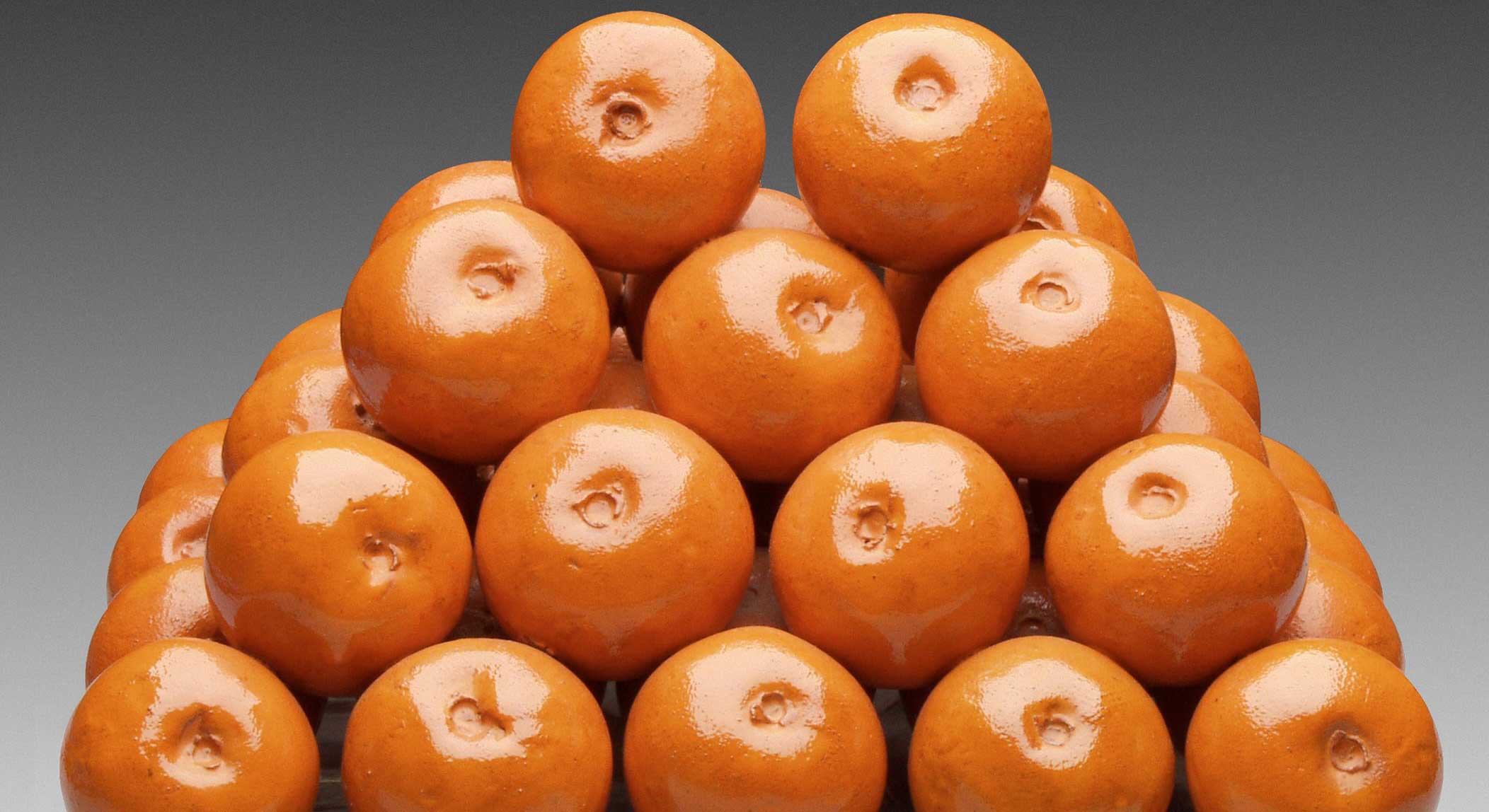 A ceramic sculpture of a triangular stack of small oranges sits on a flat grey shape against a grey background.
