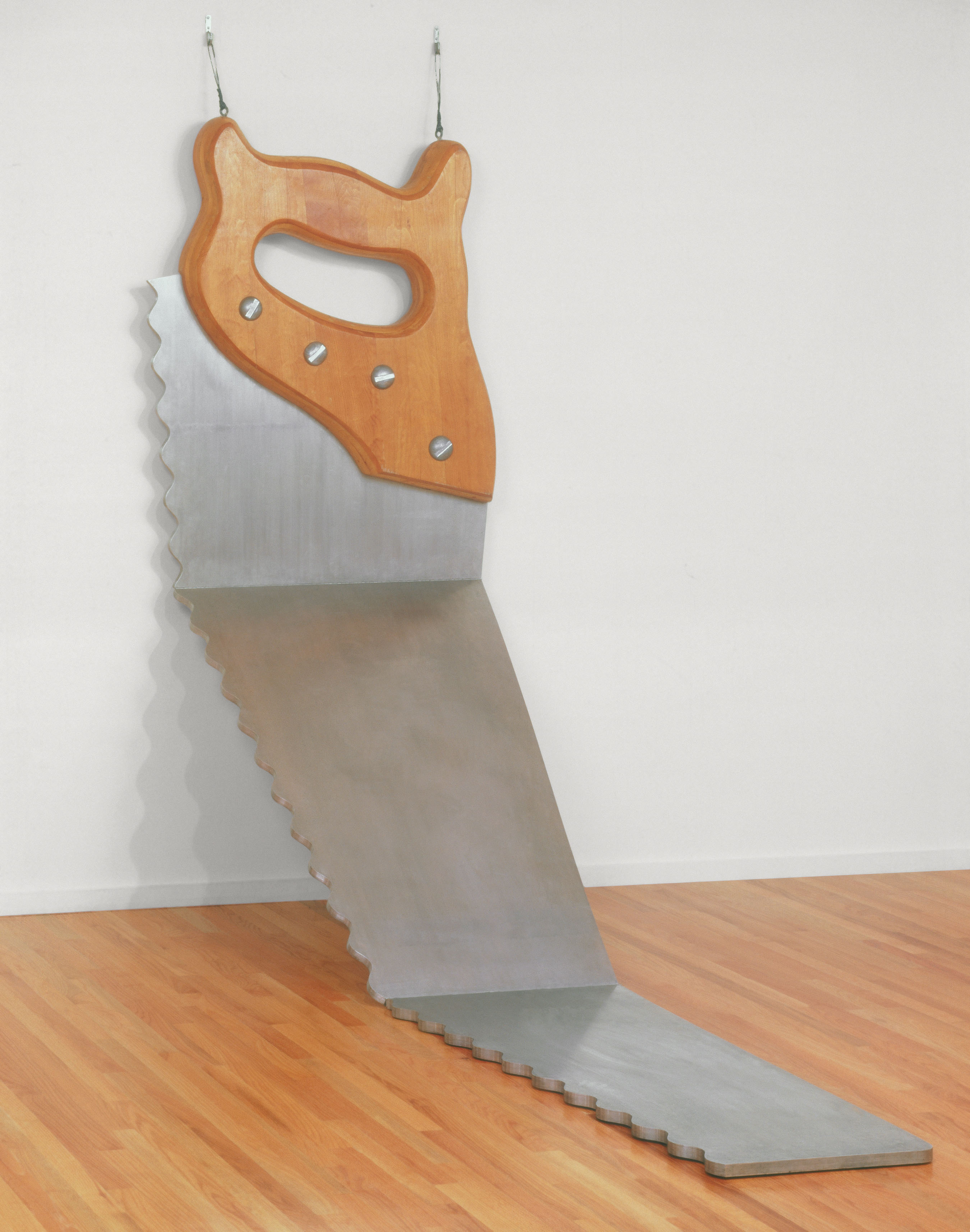 An abstract piece of artwork depicts a silver saw with a wooden handle hanging from a white wall, the blade bent in three places and draped over a hardwood gallery floor.