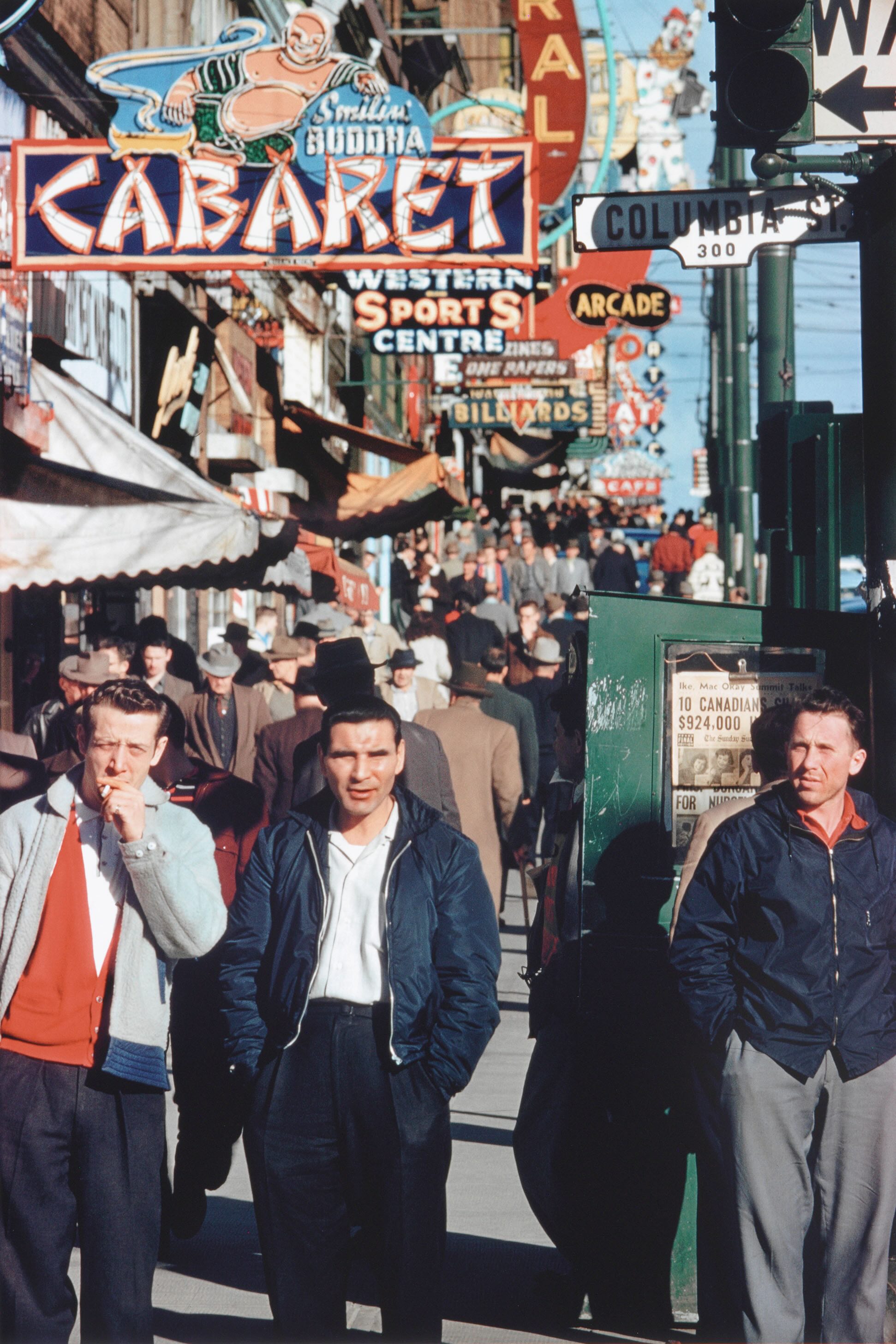 A colourful piece of 1958 street photography depicts three men in the foreground in casual streetwear. Behind them are crowds of other men walking down a busy sidewalk under storefront awnings and neon signs.  
